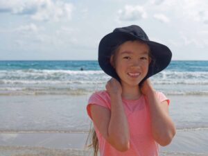A young girl stands by the water at the beach smiling at the camera.