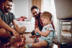Two foster dads happily watch their toddler play with building blocks on their living room floor.