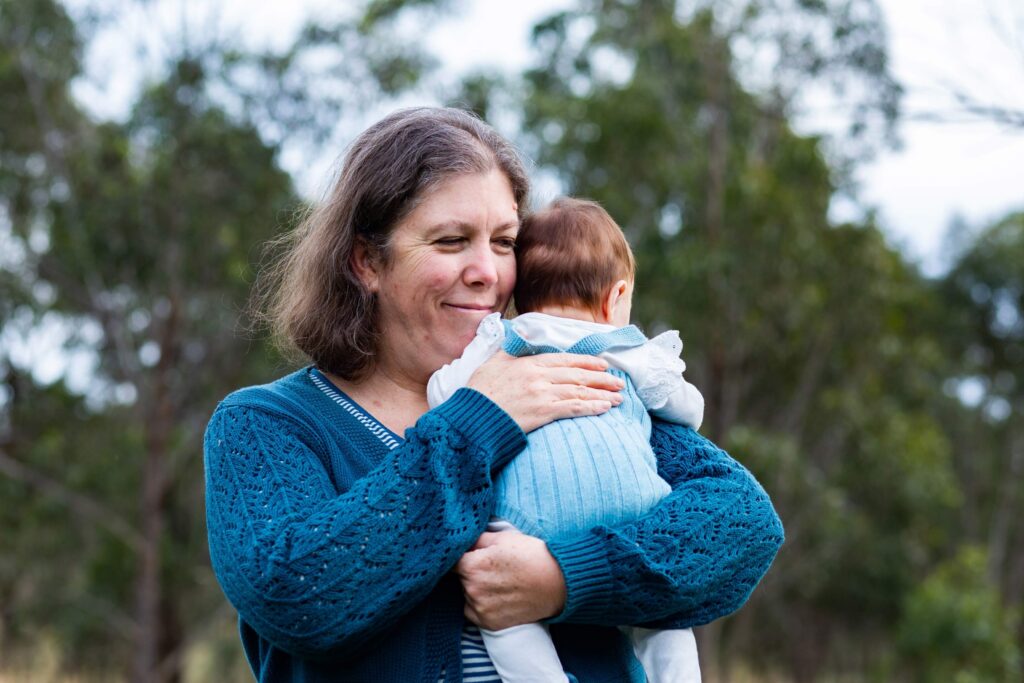 A middle-aged woman holds a baby to her chest and smiles to someone off-camera. They're in a park or bush land.